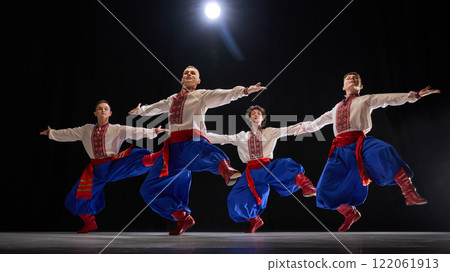 Four male dancers in formation, arms outstretched, dressed in embroidered shirts, blue trousers, and red boots, highlighting unity and tradition against black studio background. 122061913