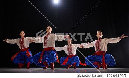 Group of male dancers performing synchronized jump, dressed in traditional embroidered shirts, blue trousers, and red boots, expressing traditions against black studio background. Group of male dancers performing synchronized jump, dressed in traditional embroidered shirts, blue trousers, and red boots, expressing traditions against black studio background. 122061915