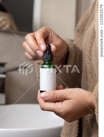Hands in brown bathrobe holding green dropper bottle with label closeup, cosmetic mockup 122062079
