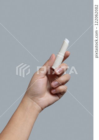 Hand holding white opened lip balm stick against grey close up, cosmetic mockup, copy space 122062082