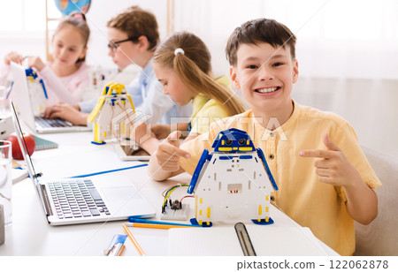 A young boy sits in a classroom, beaming with pride as he points to a robot he built. He is surrounded by other students who are also engaged in STEM activities. 122062878