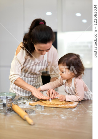 pretty mother and little child girl cutting cookies of raw gingerbread dough in kitchen pretty mother and little child girl cutting cookies of raw gingerbread dough in kitchen 122063034