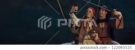 Portrait of two men, pirates in vintage costumes, holding swords and joyfully taking selfie with smartphone against dark textured background Portrait of two men, pirates in vintage costumes, holding swords and joyfully taking selfie with smartphone against dark textured background 122063523