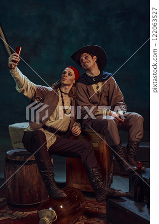 Two men, pirates in bandana and hat, sitting and taking selfie with smartphone against dark background Two men, pirates in bandana and hat, sitting and taking selfie with smartphone against dark background 122063527