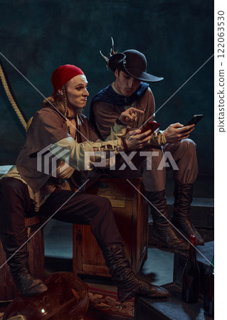 Two young men, medieval pirates in vintage costumes sitting and watching social media feed on smartphone, discussing news against dark background in dimly-lit room Two young men, medieval pirates in vintage costumes sitting and watching social media feed on smartphone, discussing news against dark background in dimly-lit room 122063530