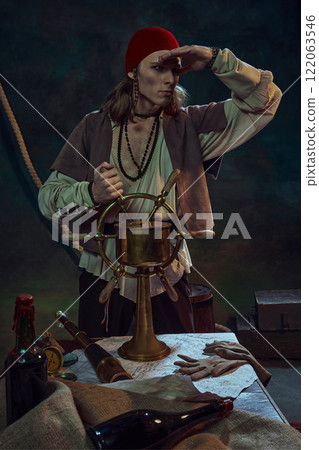 Young man, pirate in red bandana holding ship wheel, gazing into horizon with determined look against dark background with maritime tools 122063546