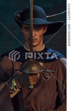 Portrait of handsome young man, pirate with dark eyes and intense gaze, in vintage costume and dark hat, holding sword next to his face 122063573