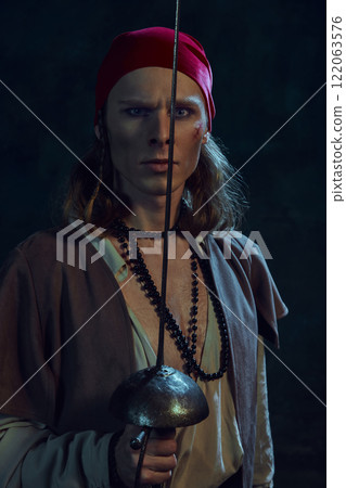 Portrait of young man, pirate with wounded face, in red bandana, retro clothes and sword next to his face, standing with intense gaze against dark background 122063576