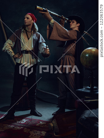 Brave pirates. Two men, dressed in vintage costumes, standing in a dark-lit rood with vintage items. One man stands with sword, while his companion looking in telescope. 122063579