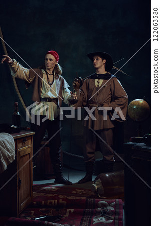 Two men dressed in historical pirate costumes, one in red bandana and necklaces, pointing forward, and other in feathered hat holding sword. 122063580