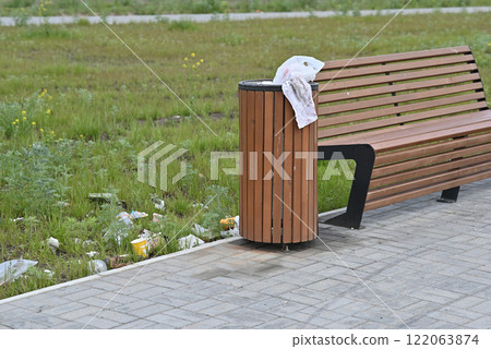 Wooden bench and trash can with litter scattered in a park setting. Wooden bench and trash can with litter scattered in a park setting. 122063874