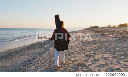 Girl Walks And Plays The Bass Instrument On The Beach Near The Ocean At Sunset Girl Walks And Plays The Bass Instrument On The Beach Near The Ocean At Sunset 122064003