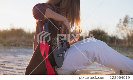 Girl Plays The Bass Instrument Sitting On The Beach At Sunset Near The Ocean 122064019
