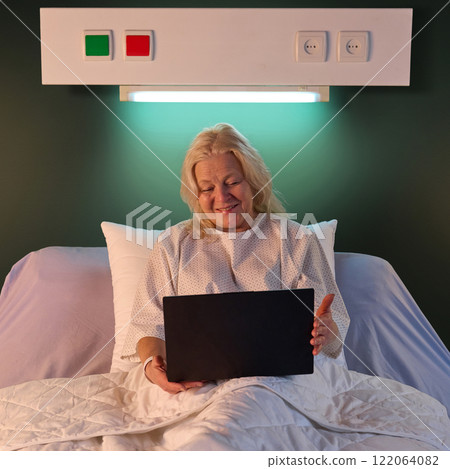 Happy smiling elderly woman sitting in hospital bed and chatting with family during online video call via laptop in the evening against green wall 122064082