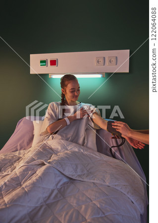 Young woman in her 20s sitting in hospital bed while hospital worker measuring her blood pressure against green wall. Modern clinic and recovery process 122064088