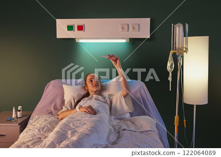 Young woman in her 20s lying in hospital bed and measuring temperature against green wall with medical equipment and pills. Modern clinic and recovery 122064089