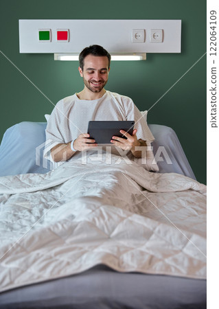 Smiling bearded man in his 40s, in hospital gown in a bed, looking on table and watching movies against green wall. 122064109