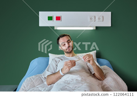 Dark-haired man in his 40s lying in hospital bed in white gown and looking on thermometer, checking temperature in hospital ward. Recovery process Dark-haired man in his 40s lying in hospital bed in white gown and looking on thermometer, checking temperature in hospital ward. Recovery process 122064111