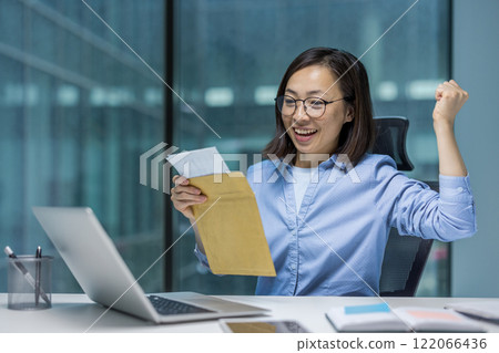 Joyful and successful Asian woman at workplace received mail notification letter envelope with good winning news, businesswoman sitting with laptop, holding hand up reading message. 122066436