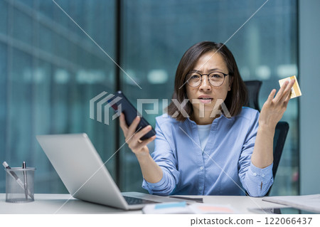 Dissatisfied and tormented Asian woman looking at camera. Office worker looking at camera holding phone and bank credit card in hands, account blocked, mistake and fraud. 122066437