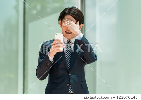 A young Asian male businessman in a suit operating a smartphone (investment, failure, mistake, shock, trouble) A young Asian male businessman in a suit operating a smartphone (investment, failure, mistake, shock, trouble) 122070145