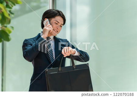 一位身穿西裝的年輕亞洲男性商人一邊用智慧型手機說話,一邊查看手錶上的時間 一位身穿西裝的年輕亞洲男性商人一邊用智慧型手機說話,一邊查看手錶上的時間 122070188