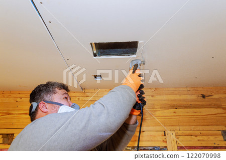 Worker is using power saw tool to cut into drywall for ventilation while wearing safety gear in new room. Worker is using power saw tool to cut into drywall for ventilation while wearing safety gear in new room. 122070998