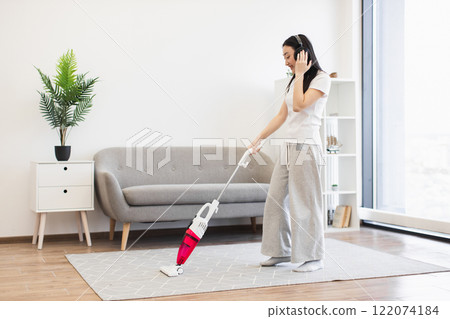 Asian female in casual attire vacuuming living room while listening to music, creating relaxed home atmosphere. 122074184
