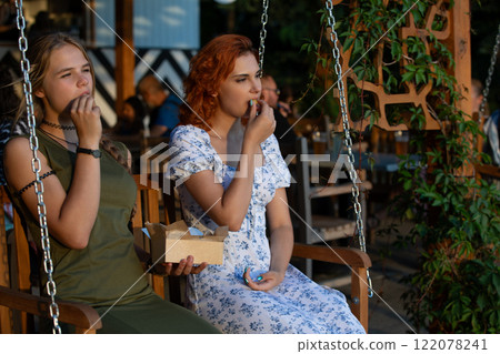 Two friends are happily enjoying snacks while swinging outdoors in a warm, green setting 122078241