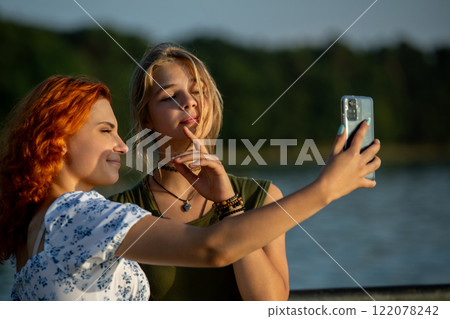 Two close friends joyfully taking a vibrant selfie together by the serene waters edge 122078242