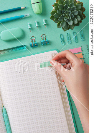 Hand sticks a post-it note in an open notebook with a blank page. Mint green stationery, pastel background. Hand sticks a post-it note in an open notebook with a blank page. Mint green stationery, pastel background. 122078259