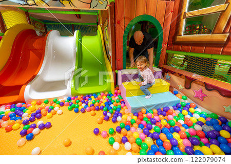 A colorful indoor playground with a ball pit, slides, and a child playing under adult supervision. A colorful indoor playground with a ball pit, slides, and a child playing under adult supervision. 122079074