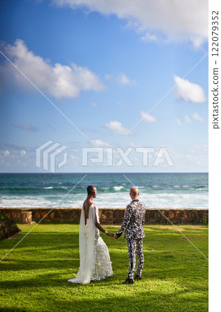 Couple at tropical villa wedding. Bride in white dress holds hands with groom in suit. Ambiguous gender fluid black people. Ocean background. LGBTQ theme. Perfect for wedding event agency travel. 122079352