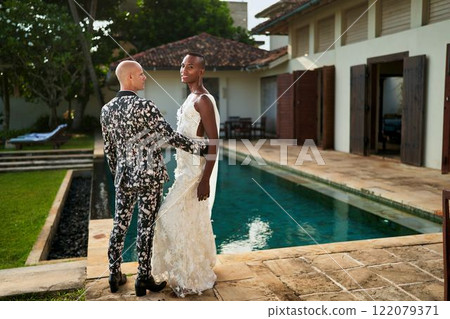 Ambiguous gender fluid person in white wedding dress beside groom in floral suit at tropical villa. Inclusive couple celebrates unique ceremony near pool. Trendy gay wedding fashion. 122079371