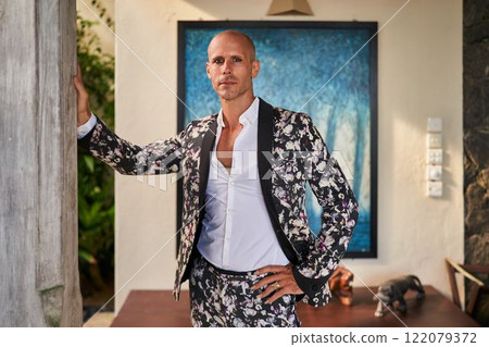 Bald man in floral suit leans on wall inside villa. Daytime setting with modern decor. Wedding attire. Calm expression suggests confident mood. Stylish groom or fashion statement theme. 122079372