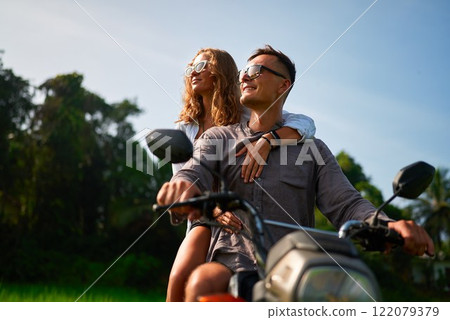 Young couple on motorbike. Smiling lovers riding through rich green jungle. Woman hugs man, both wear sunglasses. Romantic trip for adventure, love, honeymoon. Explore nature together. Young couple on motorbike. Smiling lovers riding through rich green jungle. Woman hugs man, both wear sunglasses. Romantic trip for adventure, love, honeymoon. Explore nature together. 122079379