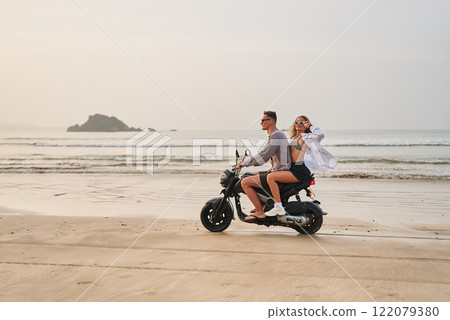 Couple rides motorbike along sandy coast. Man in sunglasses drives, woman in bikini enjoys breeze. Adventurous travel. Beach view, ocean waves, relaxed vibe. Ideal for wanderlust, freedom. 122079380