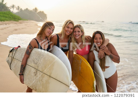 Group of four women with surfboards stand on sandy beach. Friends in swimsuits enjoy summer surf camp. Smiling and posing for photo by the sea. Tropical surfing lesson vibe. 122079391