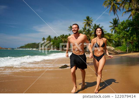 Joyful barefoot couple runs on sandy beach by ocean. Man and woman in swimwear enjoy tropical coast run. Fun, sunbathing, fitness beachside. Cheerful duo exercise, laugh near sea on sunny day. 122079414