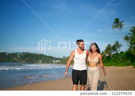 Man and woman hold hands walking on sandy beach. Couple enjoys leisure walk by sea. Smiling young partners near ocean. Romantic vacation stroll on shore. Male, female in casual beachwear together. 122079429
