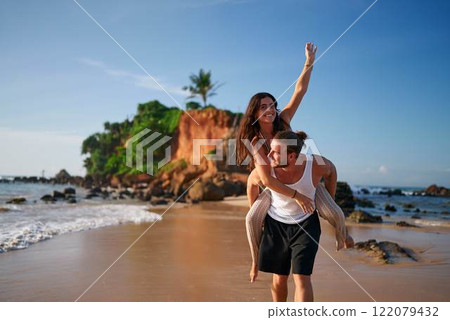 Joyful couple enjoys piggyback ride on sandy beach during sunset. Man carries girlfriend on back, both smiling, near tropical sea. Romantic vacation, playful moments by ocean, island getaway. Joyful couple enjoys piggyback ride on sandy beach during sunset. Man carries girlfriend on back, both smiling, near tropical sea. Romantic vacation, playful moments by ocean, island getaway. 122079432