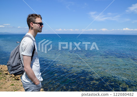 Young Russian man in sunglasses enjoying the view of the Sea of Japan on the Far East coast of Russia. Clear blue sky and calm water create a tranquil atmosphere Young Russian man in sunglasses enjoying the view of the Sea of Japan on the Far East coast of Russia. Clear blue sky and calm water create a tranquil atmosphere 122080482