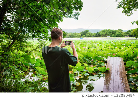 A Russian man photographing Komarov lotus lake in Duboviy Klyuch village, Primorsky Krai, Russia. Pink lotuses bloom, and a wooden pier extends into the water A Russian man photographing Komarov lotus lake in Duboviy Klyuch village, Primorsky Krai, Russia. Pink lotuses bloom, and a wooden pier extends into the water 122080494