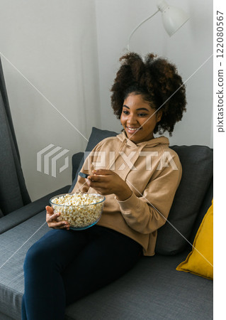 Young african american woman with a remote control in one hand and popcorn watches a television program. Cheerful enjoyment of leisure time. Streaming service, film and tv show concept. Young african american woman with a remote control in one hand and popcorn watches a television program. Cheerful enjoyment of leisure time. Streaming service, film and tv show concept. 122080567
