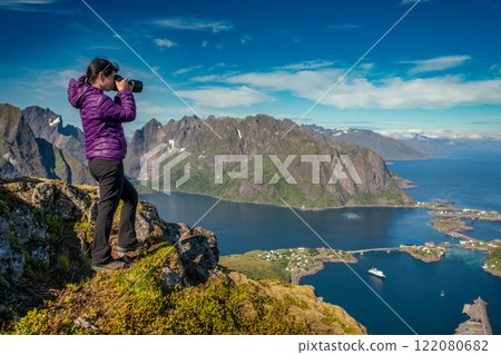 Nature photographer tourist with camera shoots while standing on top of the mountain. Amazing landscape of the Lofoten Islands from the top of Reinebringen Mountain with blue sky, Norway 122080682