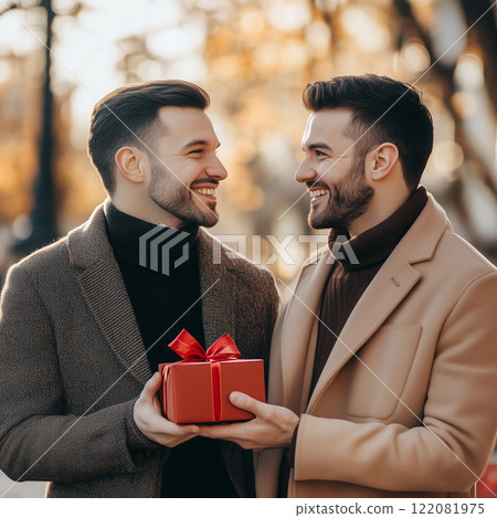 a couple of men, one man gives the other a gift in a red box with a bow outdoors a couple of men, one man gives the other a gift in a red box with a bow outdoors 122081975
