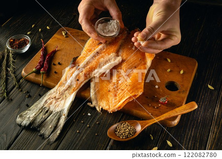 Hands skillfully season a fresh piece of smoked salmon with salt and spices in a rustic kitchen. The warm ambiance invites culinary creativity and passion for gourmet cooking Hands skillfully season a fresh piece of smoked salmon with salt and spices in a rustic kitchen. The warm ambiance invites culinary creativity and passion for gourmet cooking 122082780