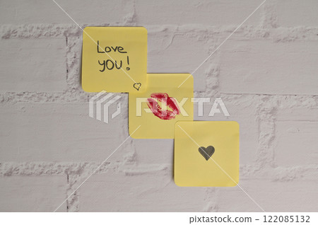 Three yellow sticker papers with lipstick kiss mark, inscription "Love you!" and small black heart on white brick wall background. Romantic message for Valentine's Day 122085132