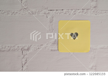 Yellow sticker paper with small black heart mark in the side on white brick wall background. Romantic message for Valentine's Day 122085133