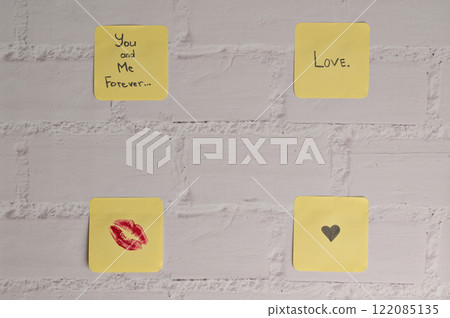 Four yellow sticker papers with lipstick kiss mark, inscription "Love you!", "You and me forever" and small black heart on white brick wall background. Romantic message for Valentine's Day 122085135